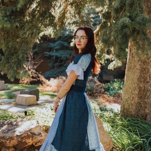 May include: A dark blue dress with white accents and a ruffled hem. The dress has short sleeves with white trim and a fitted bodice. The person is wearing beige high heels and glasses, standing in a park setting with trees and rocks.