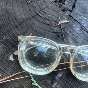 Op de afbeelding: Transparante bril met ronde glazen en een lichte tint, rustend op een donkere, getextureerde houten ondergrond. De bril heeft een klassiek ontwerp, met zichtbare scharnieren en een subtiele glans. De achtergrond bevat kleine bladeren en dennennaalden.