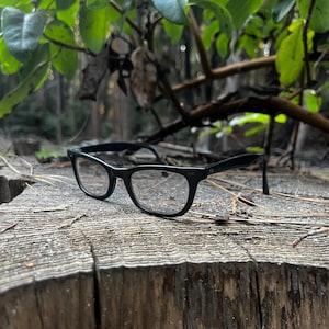 May include: Black-framed eyeglasses with clear lenses rest on a weathered wooden surface. The glasses have a classic rectangular shape and are set against a backdrop of green foliage and a natural, outdoor setting.