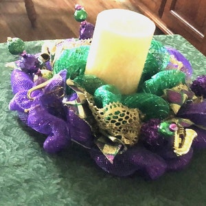 May include: A purple and green Mardi Gras wreath with a white candle in the center. The wreath is decorated with gold and purple beads, and a gold trumpet.