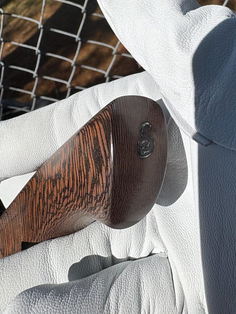 Op de afbeelding: Close-up van een golfclubkop met een donkerbruin houtnerfpatroon. De clubkop heeft een afgeronde vorm en een donker metalen embleem. De club wordt vastgehouden in een witte leren handschoen.