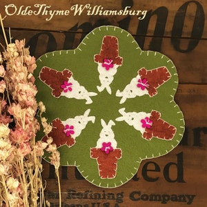 May include: A green felt Easter placemat with a scalloped edge, featuring six white bunny figures with brown flower pot hats and pink flower accents. The text "Olde Thyme Williamsburg" is at the top.