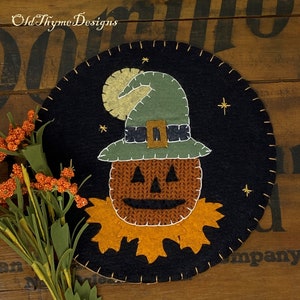 May include: A round, black felt Halloween decoration featuring a pumpkin face wearing a green witch hat with a gold buckle. The design includes a yellow moon, stars, and a stitched border. The text "Old Thyme Designs" is in the upper left corner.