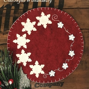 May include: A red felt round ornament with white felt snowflakes and a white stitched design of snowflakes and swirls. The ornament is on a wooden surface.