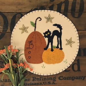 May include: A white felt circle with black stitching around the edge. The circle features a black cat, two orange pumpkins, and three white stars. The background is a light beige color.