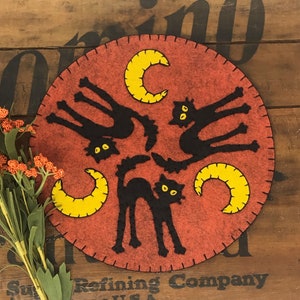 May include: A felt wall hanging with a red background and three black cats with yellow crescent moons.