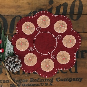 May include: A red felt Christmas ornament with a gingerbread man design. The ornament is decorated with white stitching and features eight gingerbread men in a circle.
