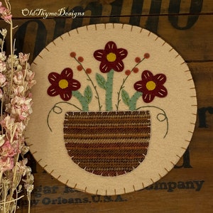May include: A felt wall hanging with a brown and white striped pot with three red flowers with yellow centers and green stems. The flowers are surrounded by small red berries.