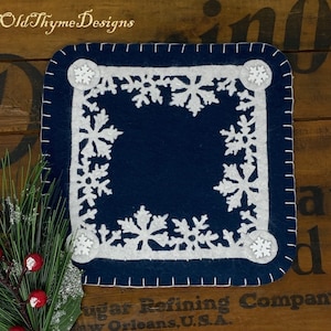 May include: A blue felt coaster with a white snowflake design. The coaster has a white border and is stitched with a white thread. The coaster is on a wooden surface.