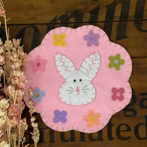 May include: A pink felt bunny rabbit applique with white felt ears and a white face. The bunny is surrounded by colorful felt flowers with yellow, pink, purple, and green centers.