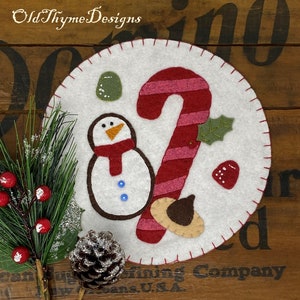 May include: A white felt circle with a red and white candy cane, a snowman, a green holly sprig, and a brown acorn. The circle is surrounded by a red stitched border. "Old Thyme Designs" is written at the top of the image.