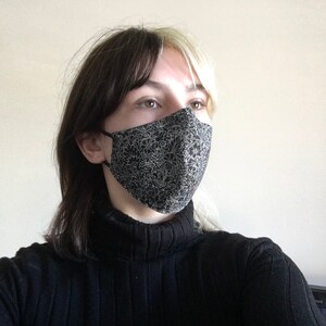 May include: A person wearing a black turtleneck sweater and a black face mask with a white floral pattern.
