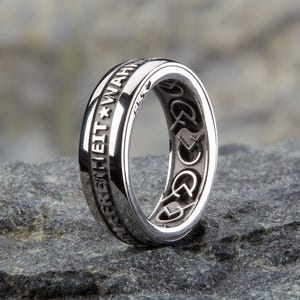 Könnte beinhalten: Ein silberner Ring mit der Inschrift "FREIHEIT * WAHRHEIT" ist um das Band graviert. Die Innenseite des Rings zeigt ein sich wiederholendes Muster aus stilisierten Symbolen.