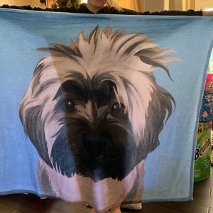 May include: A blue fleece blanket with a stylized illustration of a dog's face. The dog has brown eyes, a black nose, and white fur.