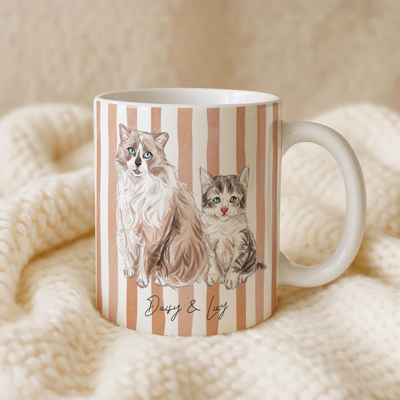 Puede incluir: Taza de cer&aacute;mica blanca con un dise&ntilde;o envolvente con dos gatos estilo acuarela, uno adulto y un gatito, sobre un fondo de rayas verticales melocot&oacute;n. La taza tiene un asa blanca y el texto "Daisy & Lucy".
