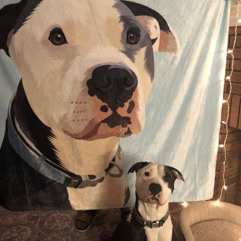 May include: A blue and white blanket with a printed image of a dog's face. A smaller dog is sitting in front of the blanket.