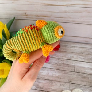 Peut inclure: Un jouet en crochet en forme de caméléon avec des rayures vertes et jaunes et des pieds orange. Le caméléon a un œil noir avec une pupille blanche et une petite corne orange sur la tête.