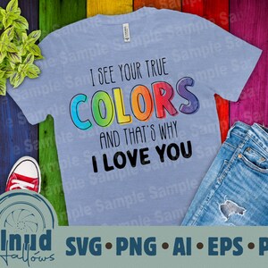 Puede incluir: Una camiseta azul claro con el texto "I see your true colors and that's why I love you" en colores arcoíris. La camiseta está sobre un fondo de madera con una planta, unos vaqueros y unos zapatos.