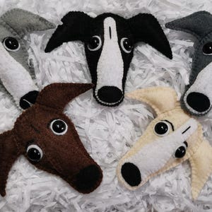 May include: Five felt dog head ornaments in various colours: grey, black and white, brown, and cream. Each has large black eyes and a white snout. The ornaments are arranged on a white, textured surface.
