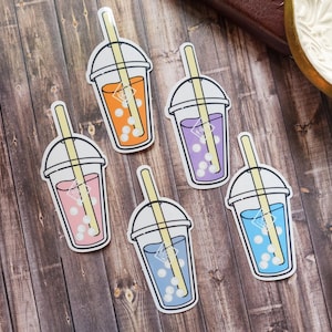 Op de afbeelding: Vijf bubble tea stickers met verschillende gekleurde drankjes. De stickers zijn op een houten oppervlak. De drankjes zijn oranje, roze, paars en blauw.