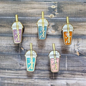 Könnte beinhalten: Fünf Emaille-Pins in Form von Bubble-Tea-Bechern mit verschiedenen Getränken und Boba-Perlen. Die Pins sind goldfarben mit weißen Deckeln und Strohhalmen.