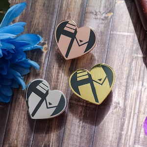 May include: Three heart-shaped pins with a black and white graphic design. The pins are in silver, gold, and rose gold.