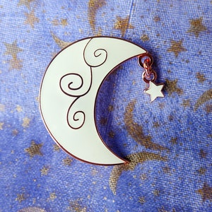May include: A white enamel crescent moon pin with a gold-tone finish and a delicate swirl design. A small star hangs from the bottom of the moon.