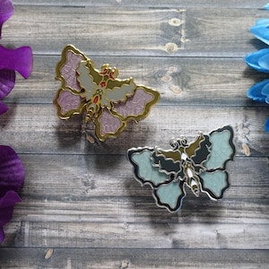 May include: Two enamel butterfly pins, one with pink and gold accents and the other with blue and silver accents. The pins are on a wooden surface.