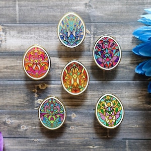 May include: Six colorful enamel pins shaped like eggs with intricate geometric designs. The pins are arranged in a circle on a wooden surface.