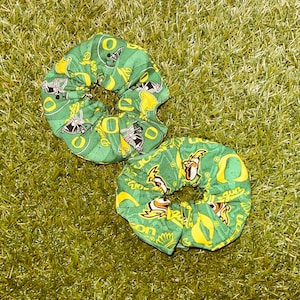 May include: Two green fabric hair scrunchies featuring a repeating pattern of yellow letter O logos, football helmets, and duck mascots. The fabric has a textured green background with the word Oregon printed in yellow throughout the design.