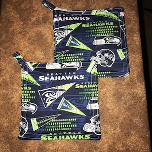 May include: Two blue and green pot holders with the Seattle Seahawks logo and text. The pot holders have a white and green geometric pattern.