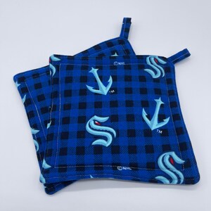 May include: Set of three blue and black checkered pot holders with the Seattle Kraken NHL logo and anchor. The pot holders have a loop for hanging.