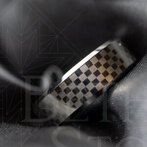 Op de afbeelding: Een zwarte en zilveren ring met een geruit patroon. De ring ligt op een zwart stoffen oppervlak.