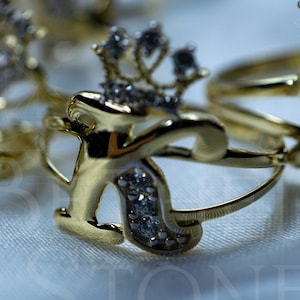 May include: Gold ring with a crown and the letter K set with clear stones. The ring is on a white fabric background.