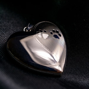 Puede incluir: Un medallón de plata en forma de corazón con un diseño de huella de pata. El medallón está sobre un fondo de tela negra.