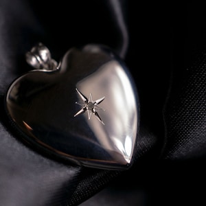 Puede incluir: Un colgante plateado en forma de corazón con un diseño de estrella, sobre tela de satén negro. El colgante tiene una superficie pulida y reflectante, y la tela crea un fondo oscuro y elegante. Joyería.