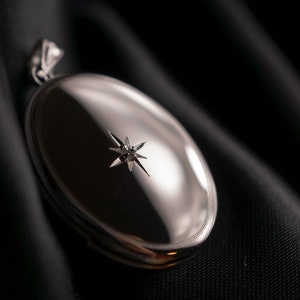 Puede incluir: Un medallón ovalado de plata con una sola piedra en forma de diamante en el centro. El medallón está sobre un fondo de tela negro.