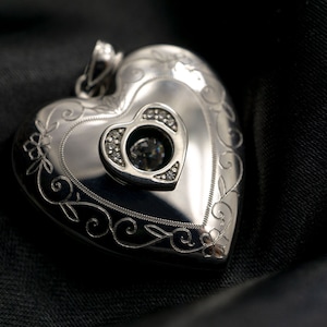 Puede incluir: Un medallón de plata en forma de corazón con un diseño intrincado y un pequeño diamante en el centro.