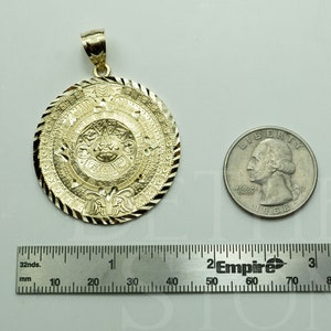 May include: A gold-coloured pendant featuring a detailed Aztec calendar design. The pendant is approximately 3.8 cm in diameter and is shown next to a US quarter for size comparison.