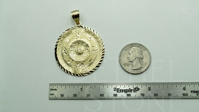 May include: A gold-coloured pendant featuring a detailed Aztec calendar design. The pendant is approximately 3.8 cm in diameter and is shown next to a US quarter for size comparison.