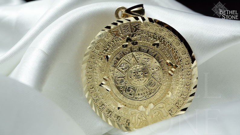 May include: A gold-coloured pendant featuring a detailed depiction of the Aztec calendar, known as the Sun Stone. The pendant is round and has a textured, ornate design.