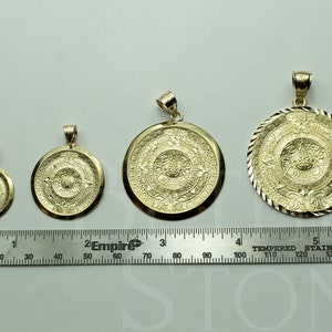 May include: Four gold-tone Aztec calendar pendants of varying sizes, displayed on a white surface with a ruler for scale. The pendants are circular and feature intricate designs.