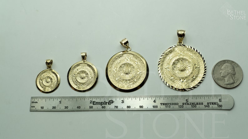 May include: Four gold-tone Aztec calendar pendants of varying sizes, displayed on a white surface with a ruler for scale. The pendants are circular and feature intricate designs.