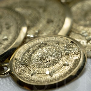 May include: A gold pendant featuring an intricate Aztec calendar design. The pendant is displayed on a white surface.