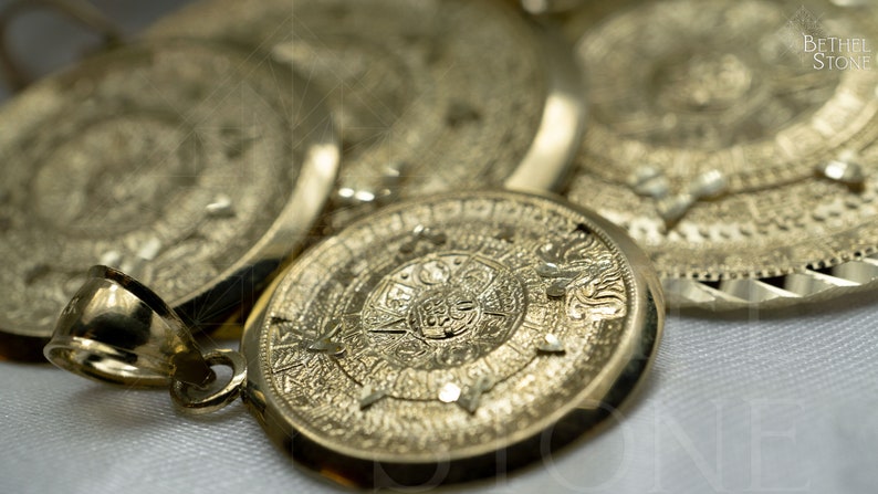 May include: A gold pendant featuring an intricate Aztec calendar design. The pendant is displayed on a white surface.