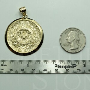 May include: A gold-coloured pendant featuring a detailed depiction of the Aztec calendar, the Sun Stone, with intricate carvings. The pendant is shown next to a US quarter for size comparison.