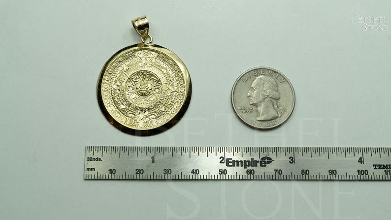 May include: A gold-coloured pendant featuring a detailed depiction of the Aztec calendar, the Sun Stone, with intricate carvings. The pendant is shown next to a US quarter for size comparison.