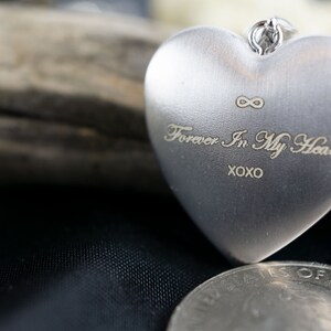 Op de afbeelding: Een zilveren hartvormig medaillon met een oneindigheidssymbool en de inscriptie "Forever In My Heart XOXO".