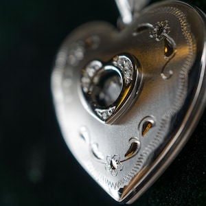 Puede incluir: Un medallón de plata en forma de corazón con una abertura más pequeña en forma de corazón en el centro. La abertura está rodeada de pequeños diamantes. El medallón tiene un diseño intrincado de flores y espirales.