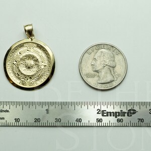 May include: A gold pendant featuring a detailed depiction of the Aztec calendar, the Sun Stone, with intricate carvings. The pendant is shown next to a US quarter for size comparison.